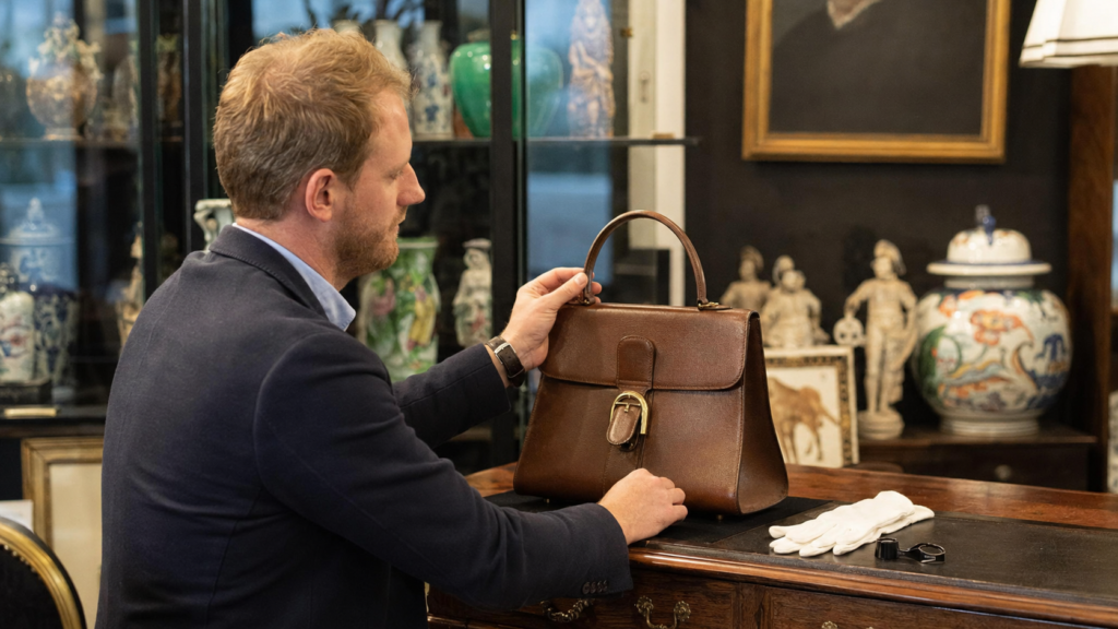 Régis Baert examine de profil un sac à main vintage en cuir brun dans une galerie d’antiquités.